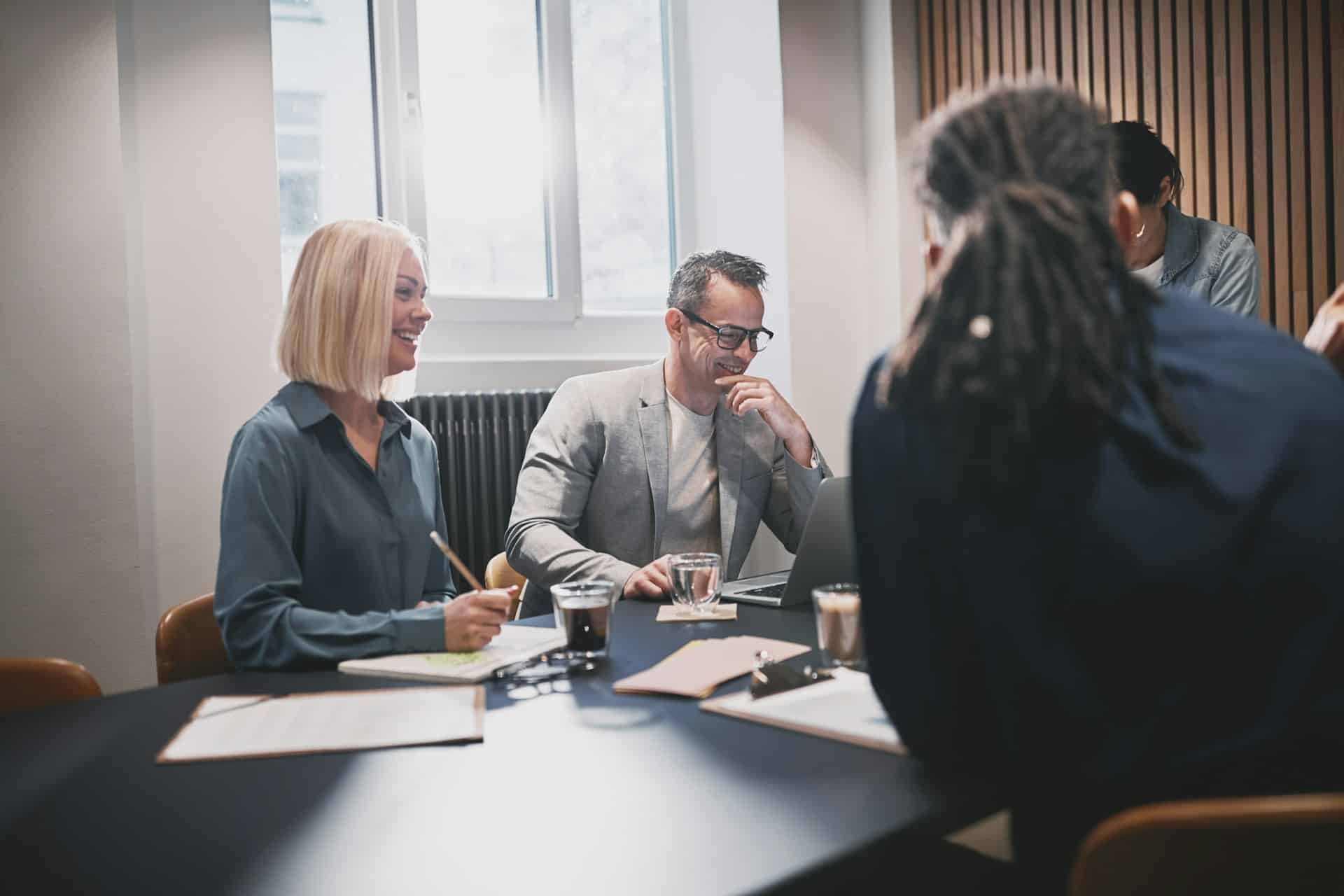 Four people are gathered around a meeting table in a boardroom, while a man smiles down at his laptop and a woman is also smiling, making notes on the pad in front of her, representing mediation in commercial disputes.