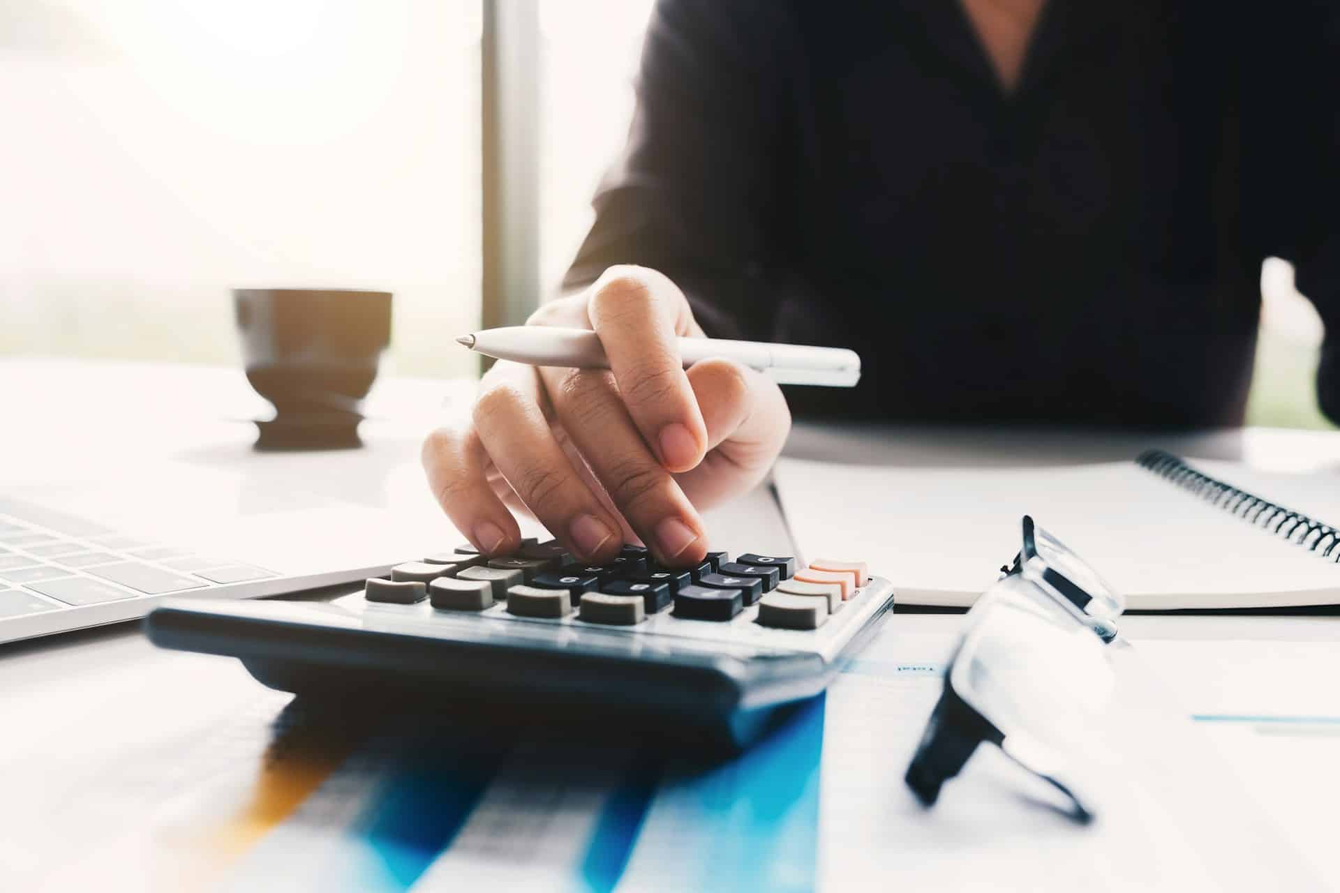 A person uses an adding machine on the desk in front of them while they record notes in a spiral notebook.