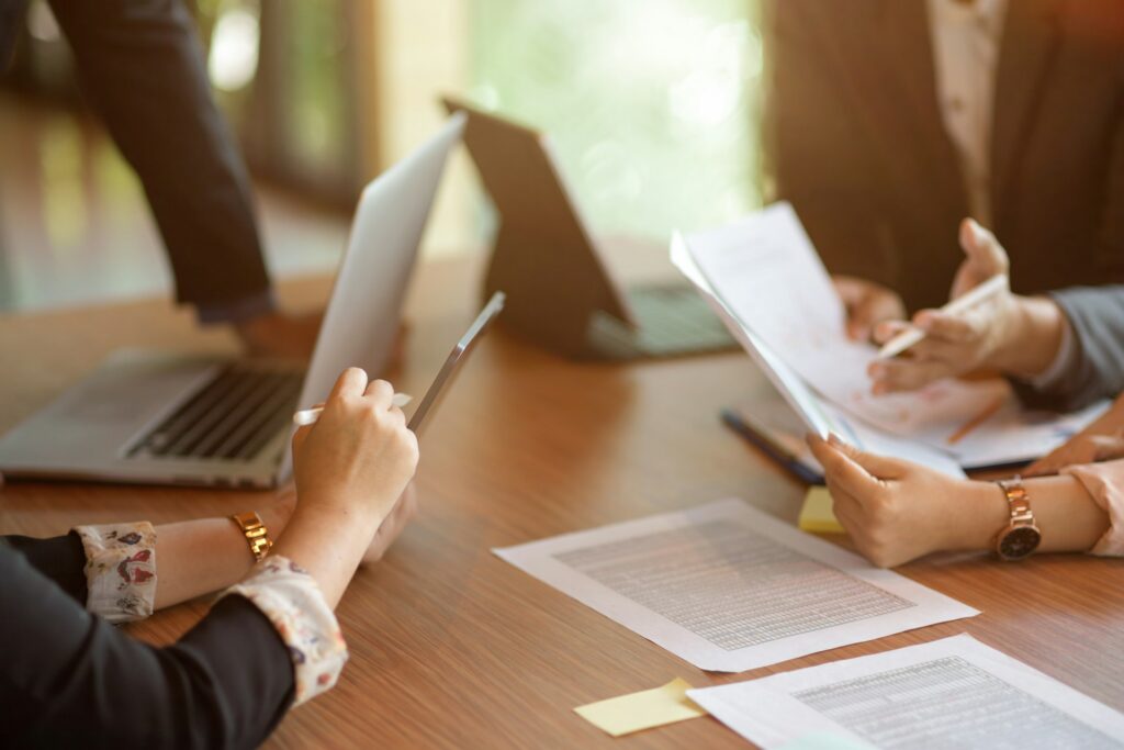 Professionals gather around a wooden meeting table and review documents in their hands and on the table as well as on the two laptops, representing mediation in supply chain and vendor disputes.