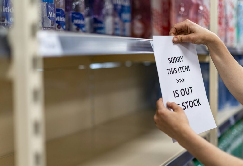 A grocery store worker, showing only their hands and forearms, hangs an 'Out of Stock' sign on an empty shelf, representing product recalls in Canada.