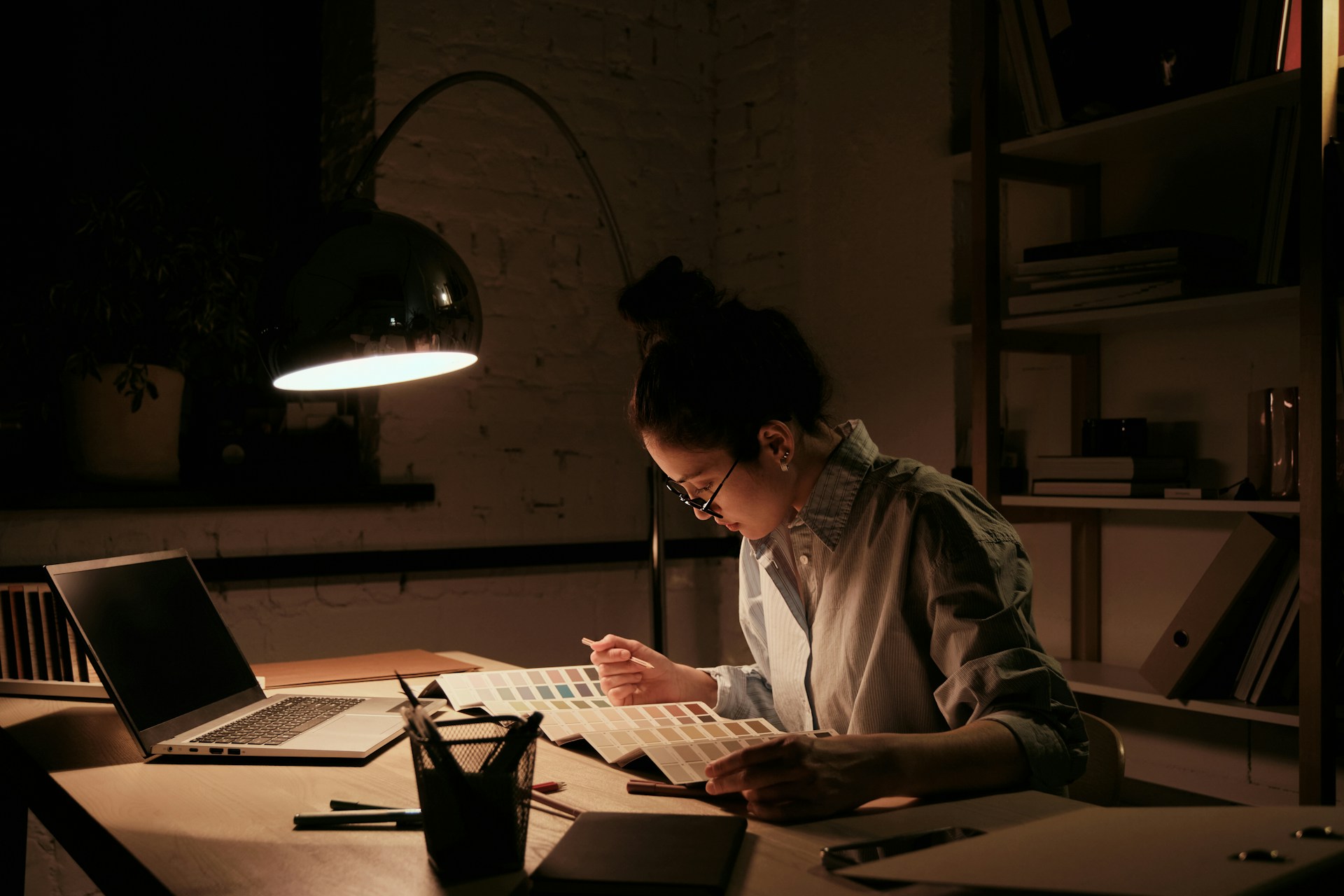 A woman works at her desk with a floor lamp illuminating her papers and laptop, and the window to outside is dark, representing on-call and after-hours work.